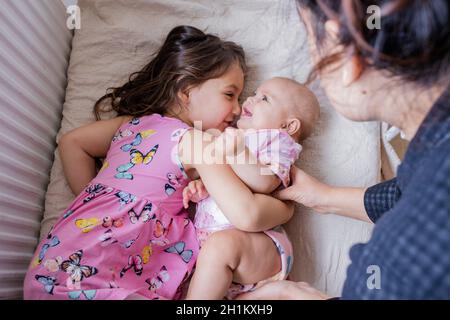 Fröhliches kleines Mädchen, das lächelnd auf dem Bett liegt und ihre glückliche Schwester neben ihrer Mutter umarmt. Zwei schöne Schwestern in rosa Mäntel liegen auf dem Bett. Stockfoto