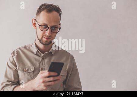 Fröhlicher kaukasischer Mann mit beigefarbenem Hemd und Brille mit Handy auf grauem Hintergrund. Auf das Telefon schauen. Fokus auf Gesicht. Stockfoto