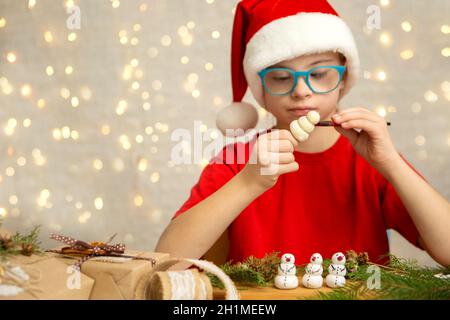 Ein Mädchen mit Down-Syndrom malt einen Schneemann aus Teig, um einen Weihnachtsbaum zu schmücken Stockfoto