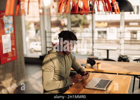 Afroamerikanischer Mann mit Laptop und Telefon in den Händen, sitzt an einem Tisch im Büro. Online arbeiten. Korrespondenz im sozialen Netzwerk Stockfoto