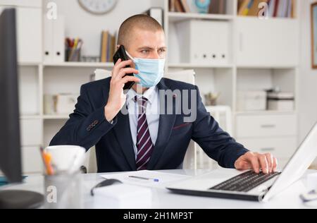 Ein männlicher Geschäftsmann in einer Schutzmaske diskutiert aktuelle Probleme mit einem Geschäftspartner am Telefon Stockfoto