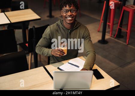 Afroamerikanischer Mann trinkt Kaffee oder Tee aus einer Papiertasse, druckt am Tisch auf einem Laptop. Arbeiten Sie am Tisch mit einem Laptop. Jobsuche über die Inter Stockfoto