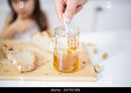 Männliche Hand pflücken Honig aus dem Glas neben einer Scheibe Brot über Holz Schneidebrett. Junges Mädchen am weißen Tisch beobachten ein Honig jar.Sweet und Licht b Stockfoto