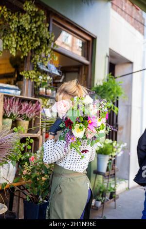 Brünette Frau hält und bedeckt ihr Gesicht hinter Blumenstrauß. Schöne und farbenfrohe Blumendarstellung vor dem Blumenladen. Houseplan Stockfoto