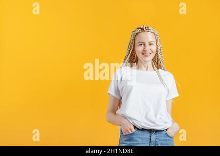 Hipper-Mode lächelndes Mädchen mit Kanekalon auf gelbem Hintergrund. Lustige, positive junge, intelligente Frau in weißem T-Shirt, lächelnd mit Spangen. Stockfoto