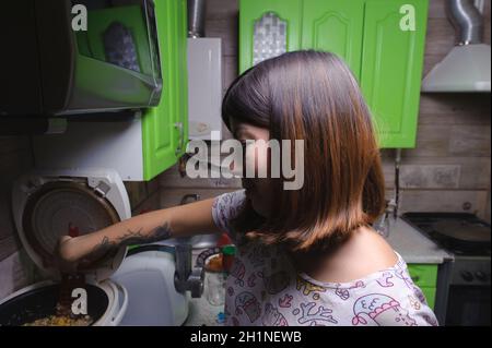 Lächelnde junge schwarzhaarige Frau, die Reis mit Gemüse mit einem Küchenschaber Spatel rührt, der für das Abendessen in einem Multikocker im k zubereitet wird Stockfoto