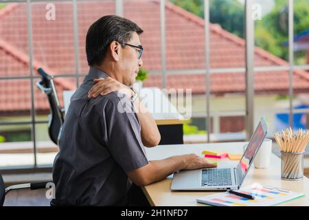Asian hart Senior Geschäftsmann arbeiten mit Laptop-Computer hat ein Problem mit Schulterschmerzen. Der alte Mann hatte Schmerzen, nachdem er lange Zeit am Schreibtisch gesessen hatte, Hea Stockfoto