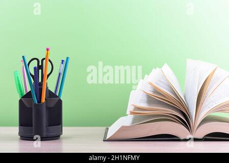 Ein Buch und ein Ständer mit Stiften, Bleistiften und Scheren auf einem hellen Holztisch. Das Buch ist geöffnet, die Blätter der Seite sind ausgefächert, ein grüner Hintergrund Stockfoto