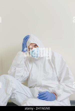 Ein trauriger Arzt in einem Schutzanzug sitzt auf dem Boden im Flur des Krankenhauses. Gesundheitspersonal während der Coronavirus-Pandemie Covid19. Stockfoto