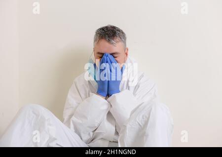Ein trauriger Arzt in einem Schutzanzug sitzt auf dem Boden im Flur des Krankenhauses. Gesundheitspersonal während der Coronavirus-Pandemie Covid19. Stockfoto