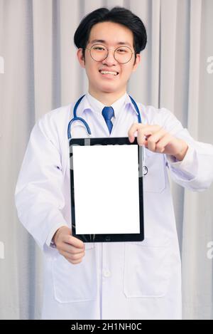 Portrait Nahaufnahme von Happy Asian junge Arzt Handsome Mann lächelnd in Uniform und Stethoskop Nackenband zeigt vor leeren Bildschirm smart digital Tisch Stockfoto