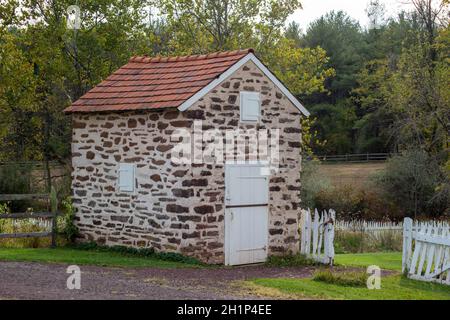 Schönes restauriertes springhouse aus Stein mit rotem Dach und weißer Holztür und Fensterläden. Malerischer weißer Holzzaun steht neben einem Tor zum g Stockfoto