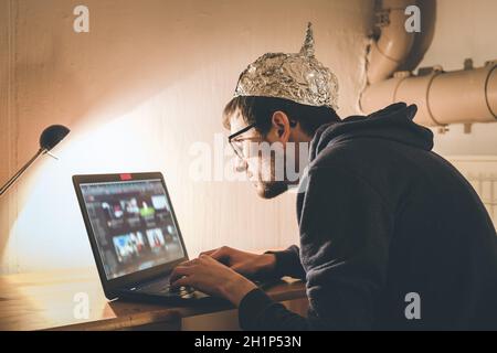 Der junge Mann mit Aluminiumkappe sitzt im dunklen Keller vor einem Laptop. Verschwörungstheorie Konzept Stockfoto