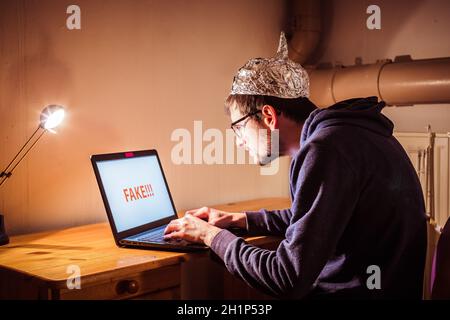 Der junge Mann mit Aluminiumkappe sitzt im dunklen Keller vor einem Laptop. Verschwörungstheorie Konzept Stockfoto