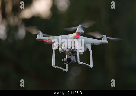 12 - Weiße Quadrotor-Drohne mit Orientierungslicht, die isoliert von dunklem unfokussiertem Hintergrund schwebt. Professionelle Kamera auf Gimbal unten gibt FPV Stockfoto