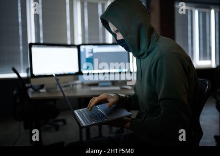 Männlicher Internet-Hacker in der Haube hält Laptop, Vorderansicht. Illegale Web-Programmierer am Arbeitsplatz, kriminelle Beschäftigung. Daten-Hacking, Cybersicherheit Stockfoto