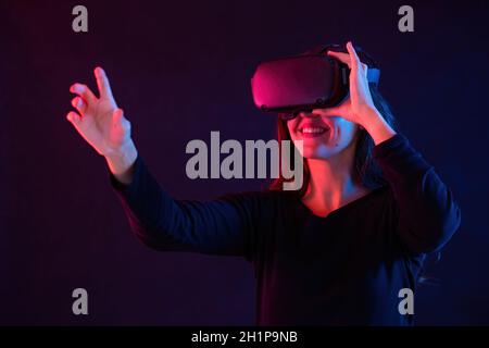 Lächelnde Frau mit Headset, die in der virtuellen Realität interagiert. Brunette mit Kopf Ausrüstung im dunklen Raum mit rotem und blauem Licht. Weibliche Berühren fictit Stockfoto