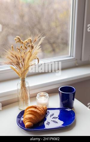 Handgefertigter blauer Tonteller mit Croissant darauf. Vase mit Weizen und Kerzen Stockfoto