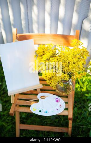 Eine Palette von Aquarell bunten Farben steht auf einem Holzstuhl und ein Strauß von gelben Blumen, kreative Hobby, Zeichnen auf der Straße. Sonniger Tag Stockfoto