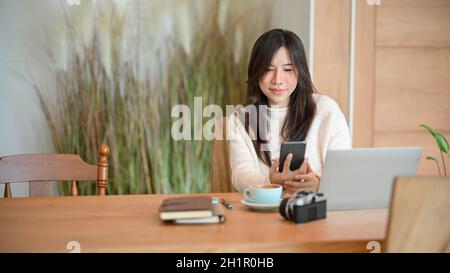 Die hübsche junge Frau sitzt in einem Café und benutzt Smartphone, Laptop, Kamera und andere Sachen auf dem Tisch. Stockfoto