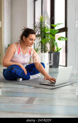 Eine Frau mittleren Alters mit Kopfhörern bereitet sich auf das Training im Internet vor. Sie sucht in den Tutorials nach Yoga- oder Pilates-Übungsvideos. Vertikales Foto. Stockfoto