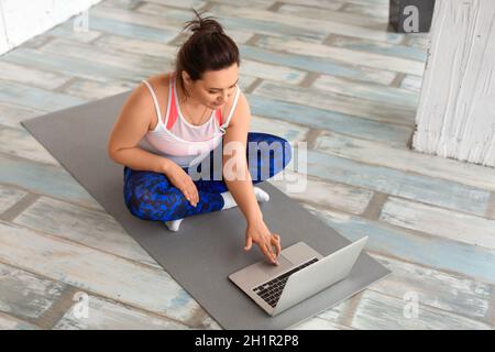 Eine Frau mittleren Alters mit Kopfhörern bereitet sich auf das Training im Internet vor. Sie sucht in den Tutorials nach Yoga- oder Pilates-Übungsvideos. Stockfoto