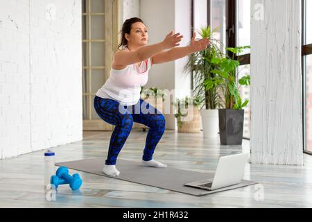 Training zu Hause - Frau mittleren Alters führt Hockübungen durch, während sie in einen Laptop-Monitor schaut. Sie nutzt ein Online-Tutorial. Stockfoto