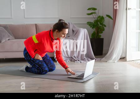 Online-Training zu Hause – eine Frau mittleren Alters mit Kopfhörern bereitet sich auf das Training im Internet vor. Sie sucht in den Tutorials nach Yoga oder Pilates Exercise V Stockfoto