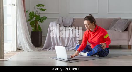 Eine Frau mittleren Alters mit Kopfhörern bereitet sich auf das Training im Internet vor. Sie sucht in den Tutorials nach Yoga- oder Pilates-Übungsvideos. Foto 2x formatieren Stockfoto