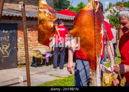 Aradac, Vojvodina, Serbien - 21. Mai 2016: Zwei rohe Schweineschinken hängen oben im Dorfhof, Metzger schneidet Fleisch. Stockfoto