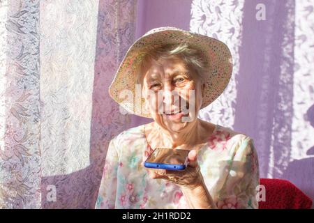 Charmante ältere Frau in rechteckiger, schwarz umrandter Brille mit Spracherkennungsfunktion auf dem Handy. Moderne ältere weibliche Holding elektronische gad Stockfoto