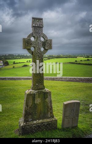 Altes keltisches Kreuz als Grabstein mit grünen Feldern und dramatischem Sturmhimmel im Hintergrund, Cashel, Irland Stockfoto