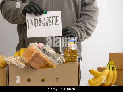 Mann hält ein Blatt Papier mit einem Schriftzug spenden und sammelt Lebensmittel, Früchte und Dinge in einem Karton, um die Bedürftigen und die Armen zu helfen, das concep Stockfoto