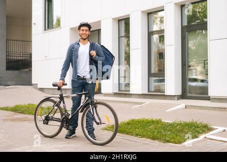 Porträt eines fröhlichen, gutaussehenden jungen, in Schutzhelm getragenem Lieferers, der in der Nähe des Fahrrads in der Stadtstraße steht und die Kamera anschaut. Stockfoto