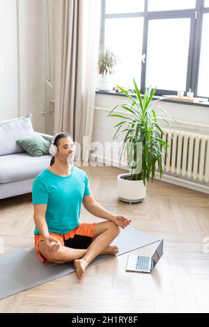 Mann mittleren Alters meditiert zu Hause - über das Internet, um das Training zu überwachen. Er hört entspannende Meditationsmusik über Kopfhörer. Home w Stockfoto