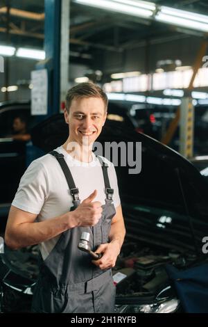 Vertikales Porträt eines lächelnden, hübschen jungen Mechanikers, der Uniform trägt und einen speziellen Schlüssel mit Ratschenschlüssel hält, der den Daumen nach oben zeigt. Stockfoto