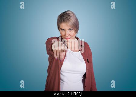 Frau mittleren Alters zeigt anklagend auf die Kamera. Blauer Hintergrund. Körpergesten und Gesichtsausdrücke Stockfoto