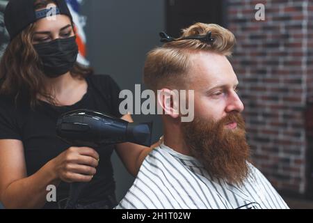 Weibliche Barbier in Maske trocknet das Haar an den Kunden mit einem Haartrockner während der sozialen Distanzierung Stockfoto