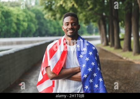 Schöner afroamerikanischer Mann mit USA-Flagge auf den Schultern, der ernsthaft auf die Kamera schaut, draußen stehend. Tag Sommer Stockfoto