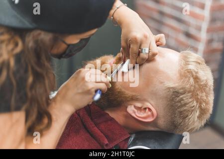 Barber Frau rasiert Bart mit einem Rasiermesser. Friseurausrüstung. Selektiver Fokus. Master in Gesichtsmaske. Stockfoto