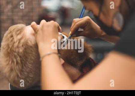 Barber Frau rasiert Bart mit einem Rasiermesser. Friseurausrüstung. Selektiver Fokus. Master in Gesichtsmaske. Stockfoto