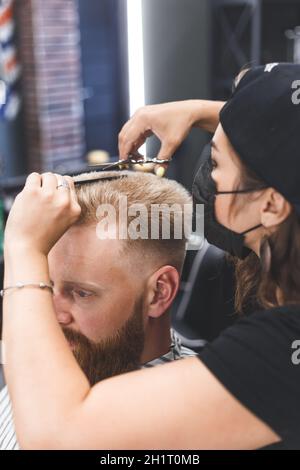 Frauen Friseur macht flache top Frisur mit Schere einen bärtigen Mann. Dienst in einer Maske von Virus Stockfoto
