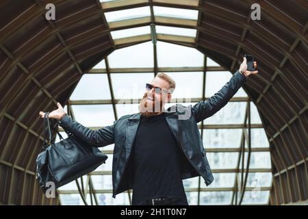 Stilvoll gekleideter junger Mann mit Bart lernt die guten atemberaubenden Nachrichten per Telefon. Lifestyle emotionales Stadtportrait. Stockfoto