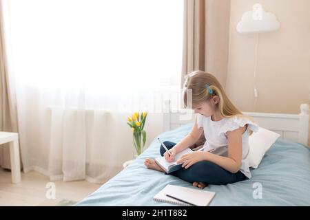 Ein kleines Mädchen sitzt auf dem Bett im Schlafzimmer und schreibt in einem blauen Buch. Bildungskonzept. Zu Hause Schulbildung. Hausaufgaben. Hochwertige Fotos Stockfoto