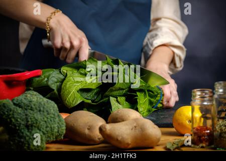 Eine Frau, die kocht, schneidet Spinat, auf dem Tisch Brokkoli, Kartoffeln, Kochtöpfe und Gewürze Stockfoto