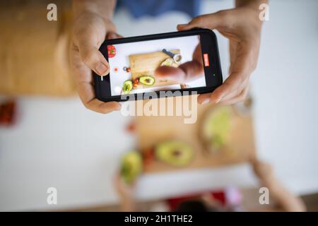 Weibliche Hände mit Smartphone Aufnahme liebenswert kleines Mädchen Verbreitung Avocado auf Scheibe Brot. Niedliches kleines Kind schneidet Gemüse auf Schneidebrett Stockfoto