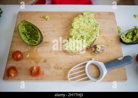 Weibliche Kinderhände verteilen ruhig Avocado auf einer Brotscheibe über dem hölzernen Schneidebrett. Yung Mädchen am weißen Tisch schneiden Gemüse. Kinder bereiten sich vor Stockfoto