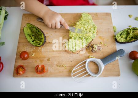 Weibliche Kinderhände verteilen Avocado auf einer Brotscheibe über dem Holzschneidebrett. Yung Mädchen am weißen Tisch schneiden Gemüse. Kinder bereiten Essen zu Stockfoto