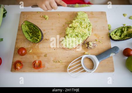 Weibliche Kinderhände verteilen Avocado auf einer Brotscheibe über dem Holzschneidebrett. Yung Mädchen am weißen Tisch schneiden Gemüse. Kinder bereiten Essen zu Stockfoto