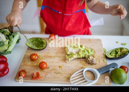 Weibliche Kinderhände verteilen Avocado auf einer Brotscheibe über dem Holzschneidebrett. Yung Mädchen am weißen Tisch schneiden Gemüse. Kinder bereiten Essen zu Stockfoto